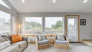 Una habitación con sofá, dos sillones de mimbre, una mesita y amplios ventanales con vistas a árboles y una terraza.