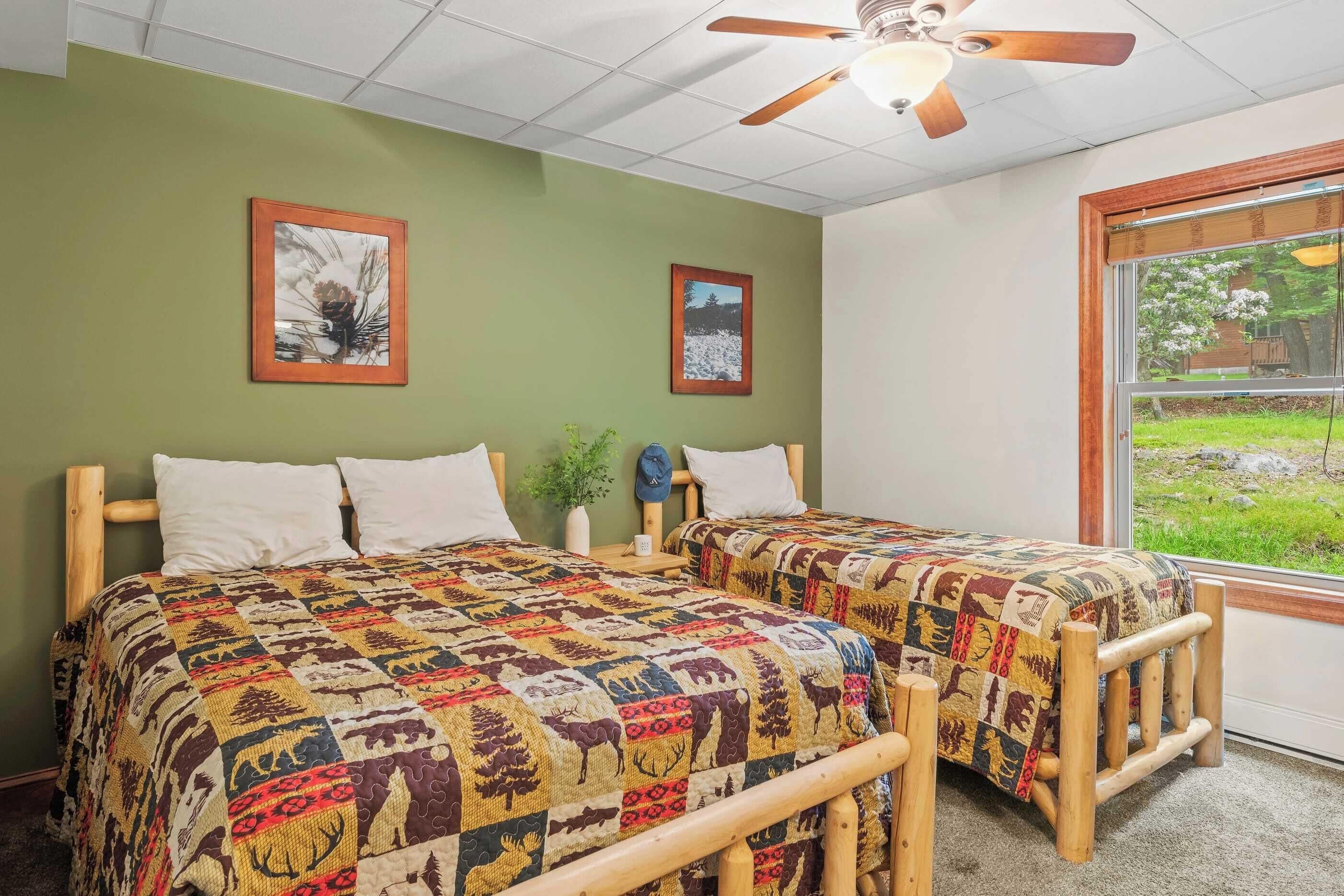 A room with two wooden beds, green walls, framed pictures, a ceiling fan, and a window with a view of greenery.