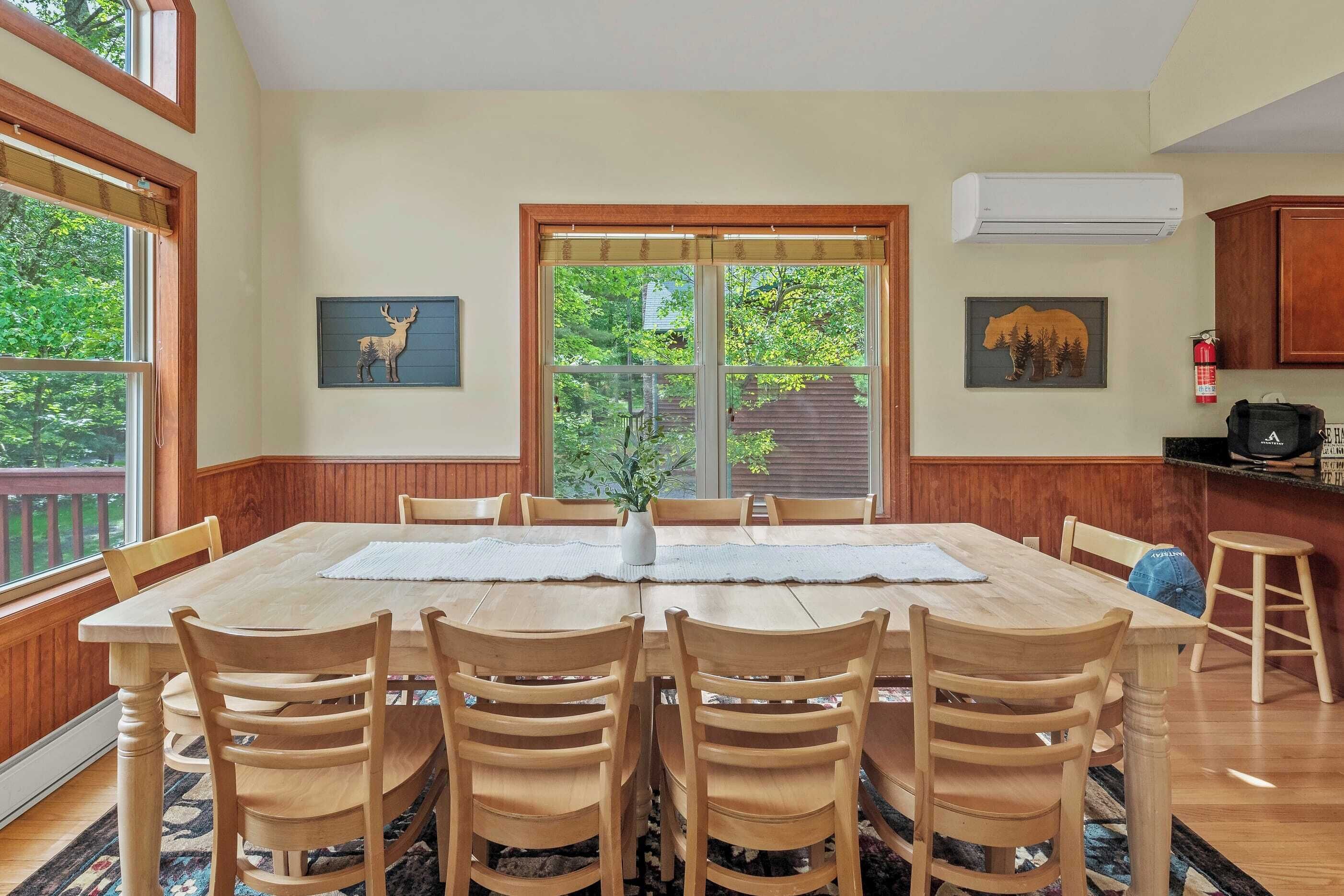 Uma sala de jantar com uma mesa e cadeiras de madeira, um vaso com flores e uma janela com vista para árvores.
