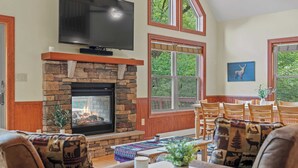 A cozy living room with a stone fireplace, a flat-screen TV mounted above it, and a large window offering a view of the outdoors.