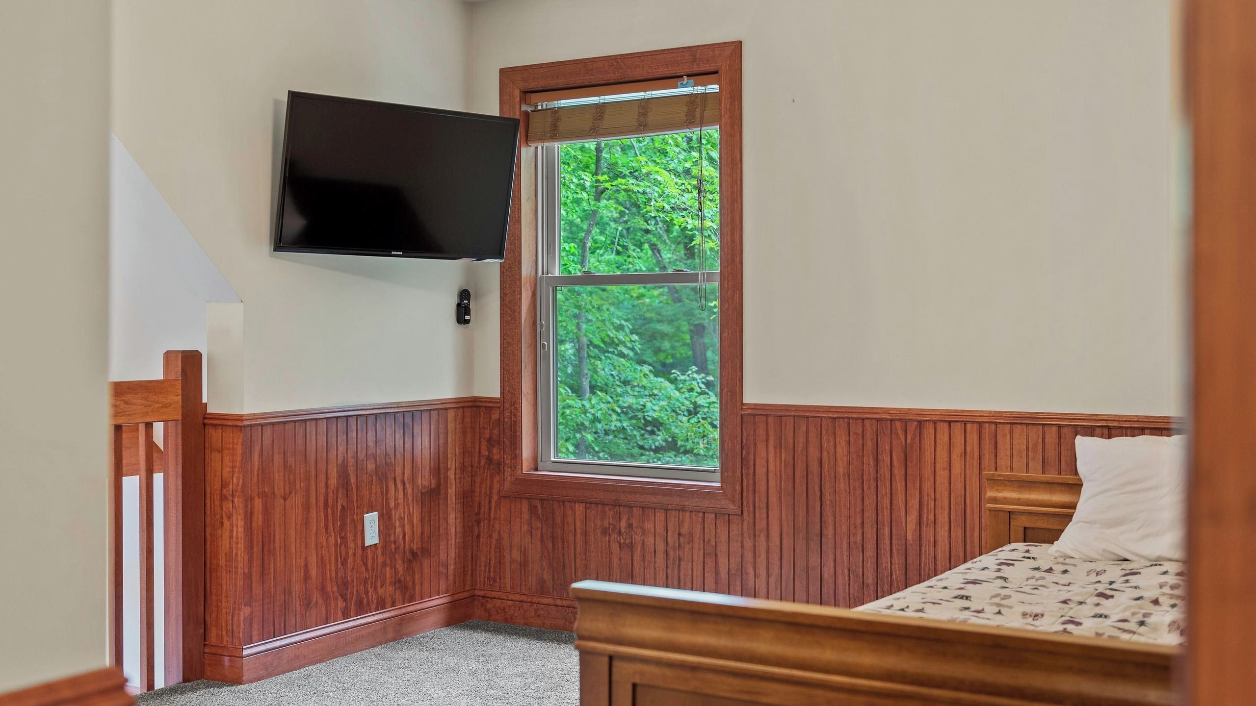 A bedroom with a wooden paneled wall, a bed with a floral bedspread, a television mounted above a window with a view of greenery, and a small bedside table.