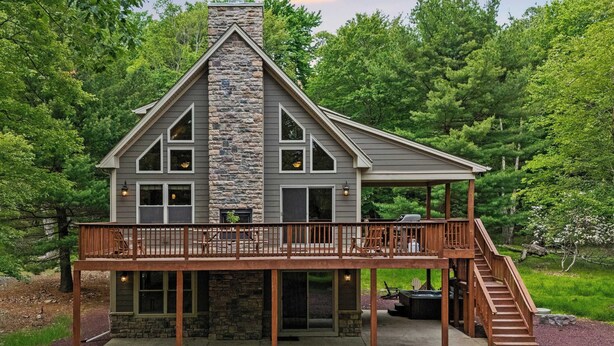 A two-story cabin with a stone chimney, a deck with a railing, and a staircase leading to the upper level.