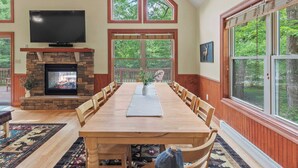 A spacious dining area with a long wooden table, stone fireplace, and large windows offering views of the outdoors.