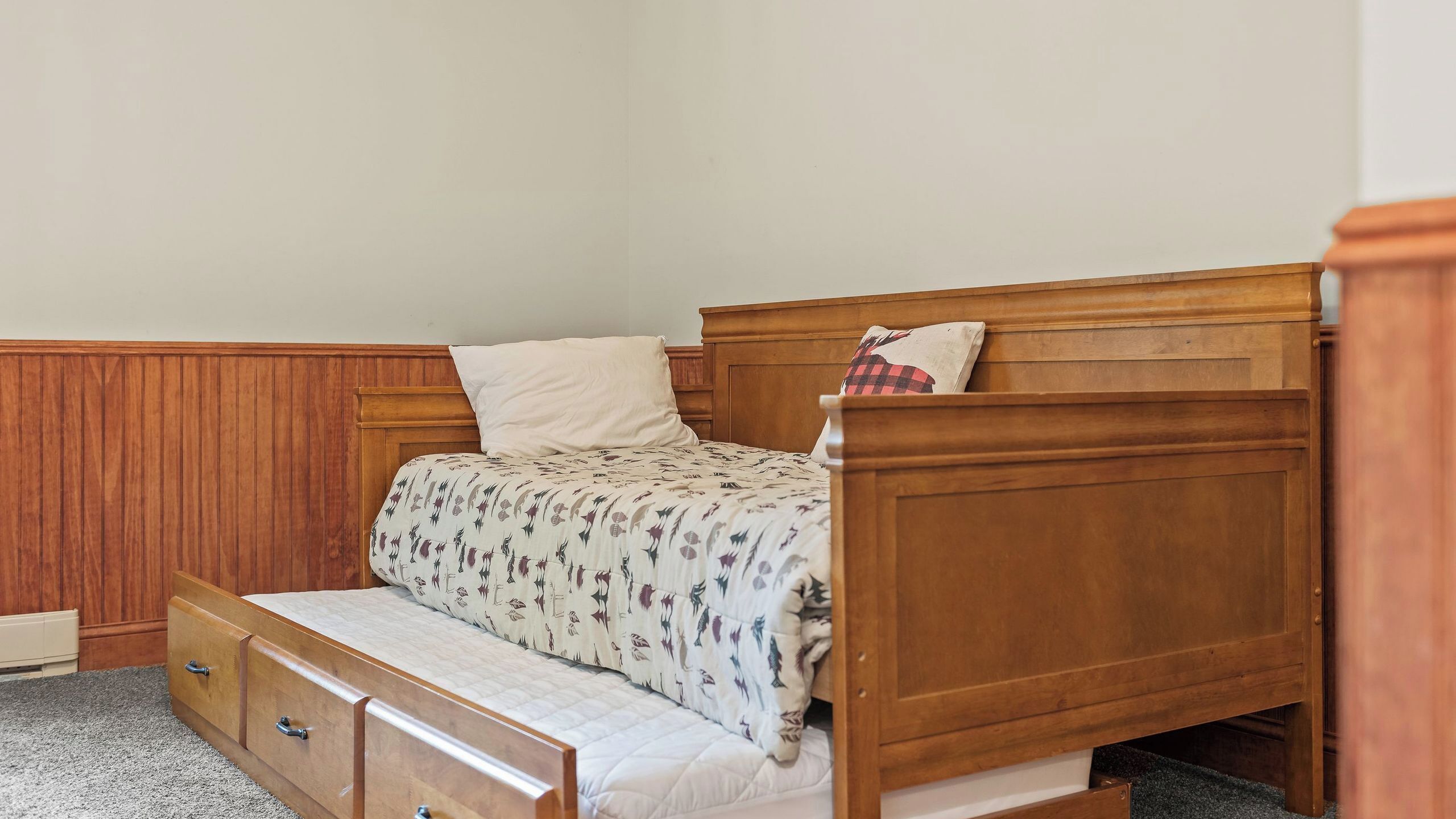 A single bed with a wooden headboard, a white pillow, a patterned bedspread, and a small bedside table with drawers.