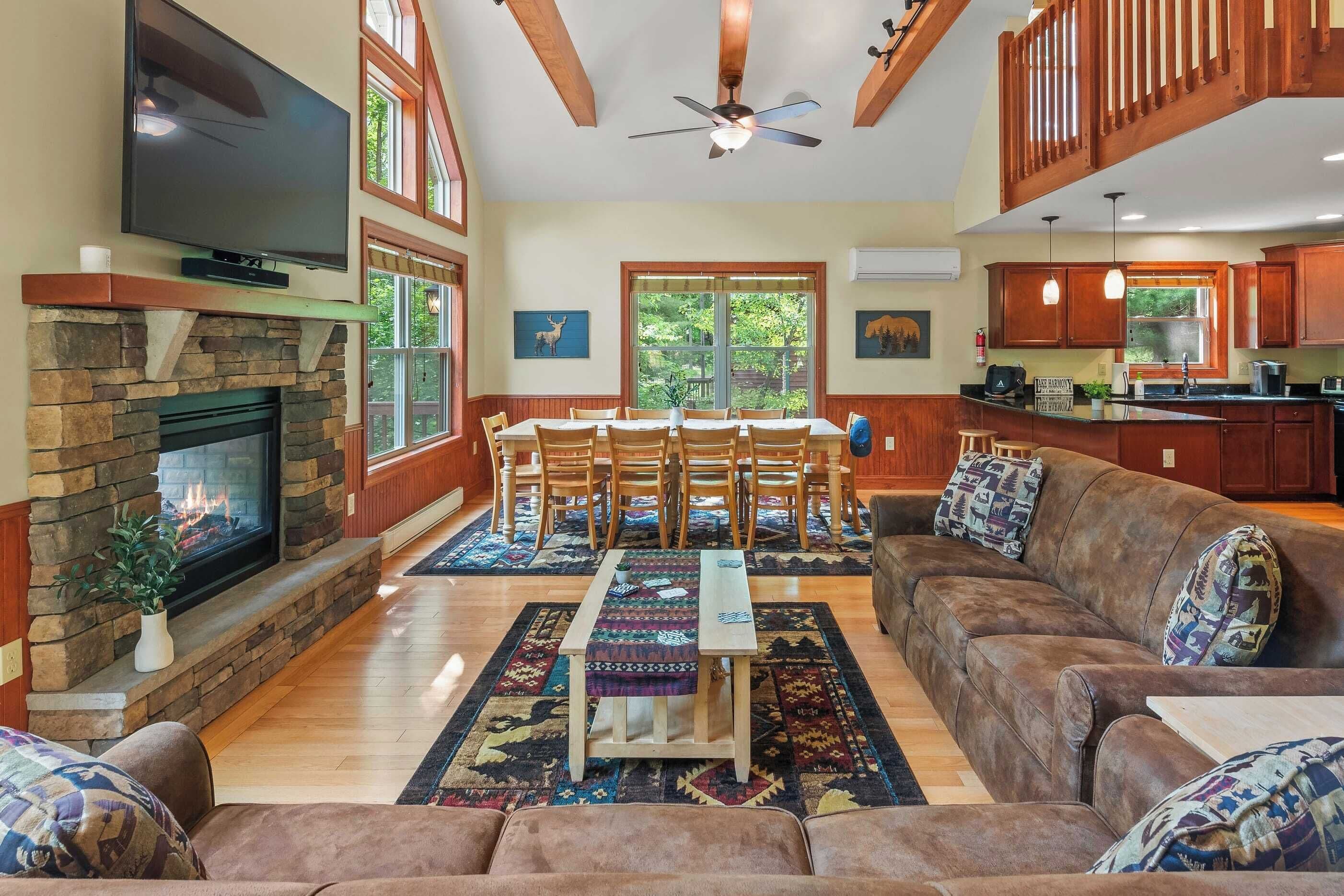 A spacious living room with a fireplace, a dining area with a wooden table and chairs, a brown leather sofa, and a kitchen area with wooden cabinets and a large window.