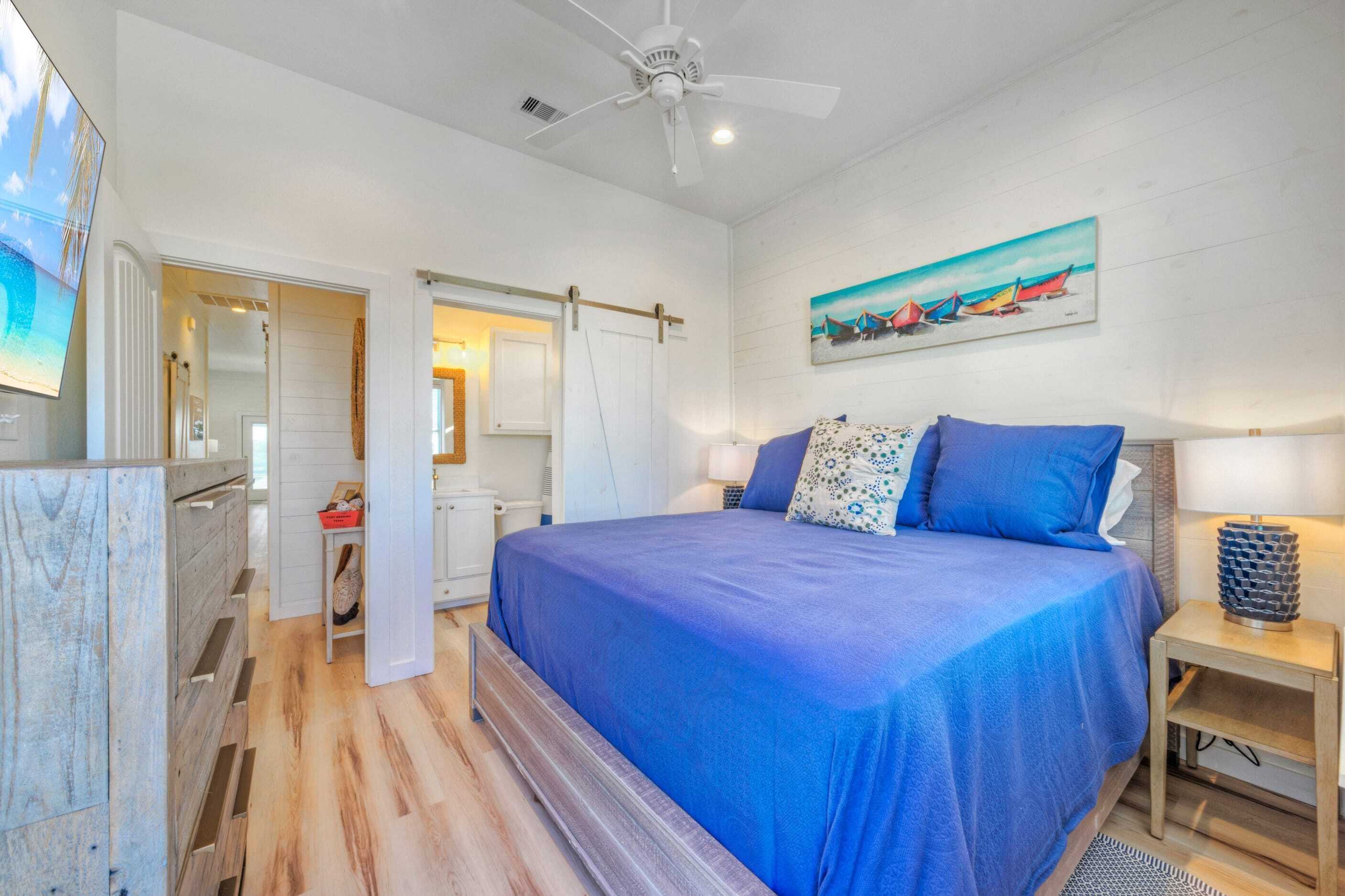 A bedroom with a large bed, a ceiling fan, a painting of boats, and a wooden dresser.