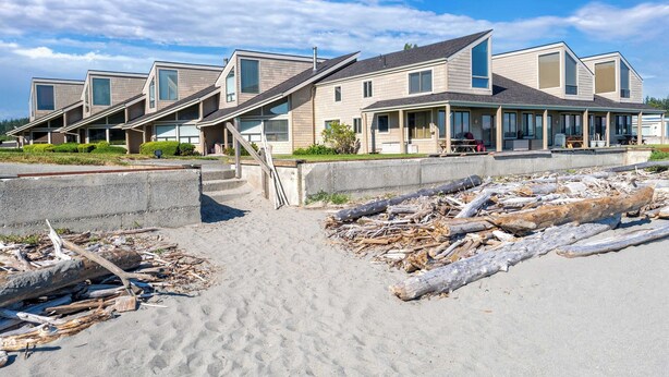 Een rij moderne strandhuizen met grote ramen, een zandstrand ervoor en een helderblauwe lucht.
