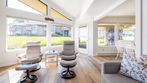 Een moderne woonkamer met een grijze bank, twee fauteuils en een eettafel. Grote ramen bieden uitzicht op een binnenplaats en omliggende gebouwen.