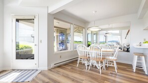 Een eetruimte met een houten vloer, witte stoelen, een ronde tafel en uitzicht door grote ramen.