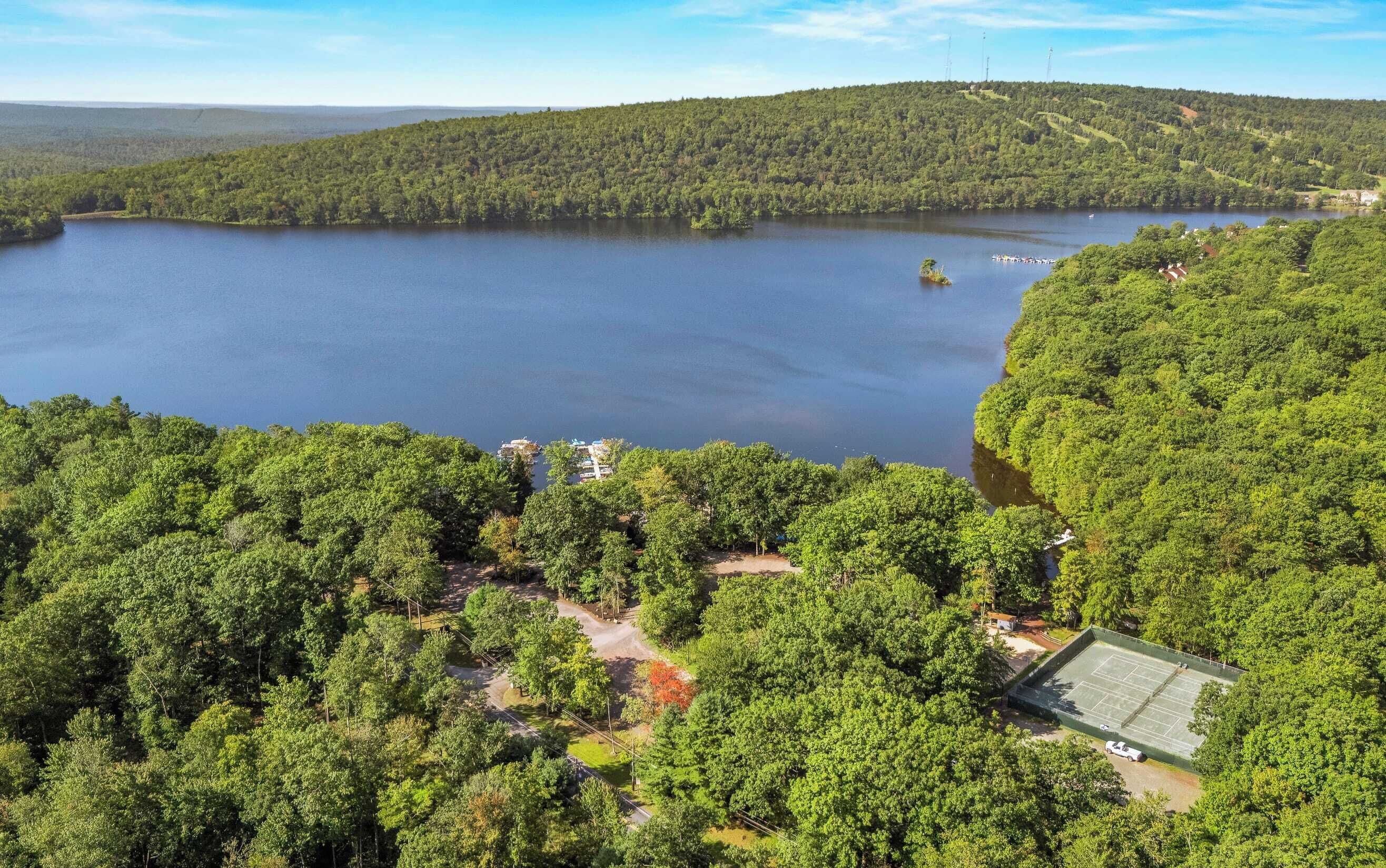 An aerial view of a lakeside property with dense forest, a tennis court, and a small dock.