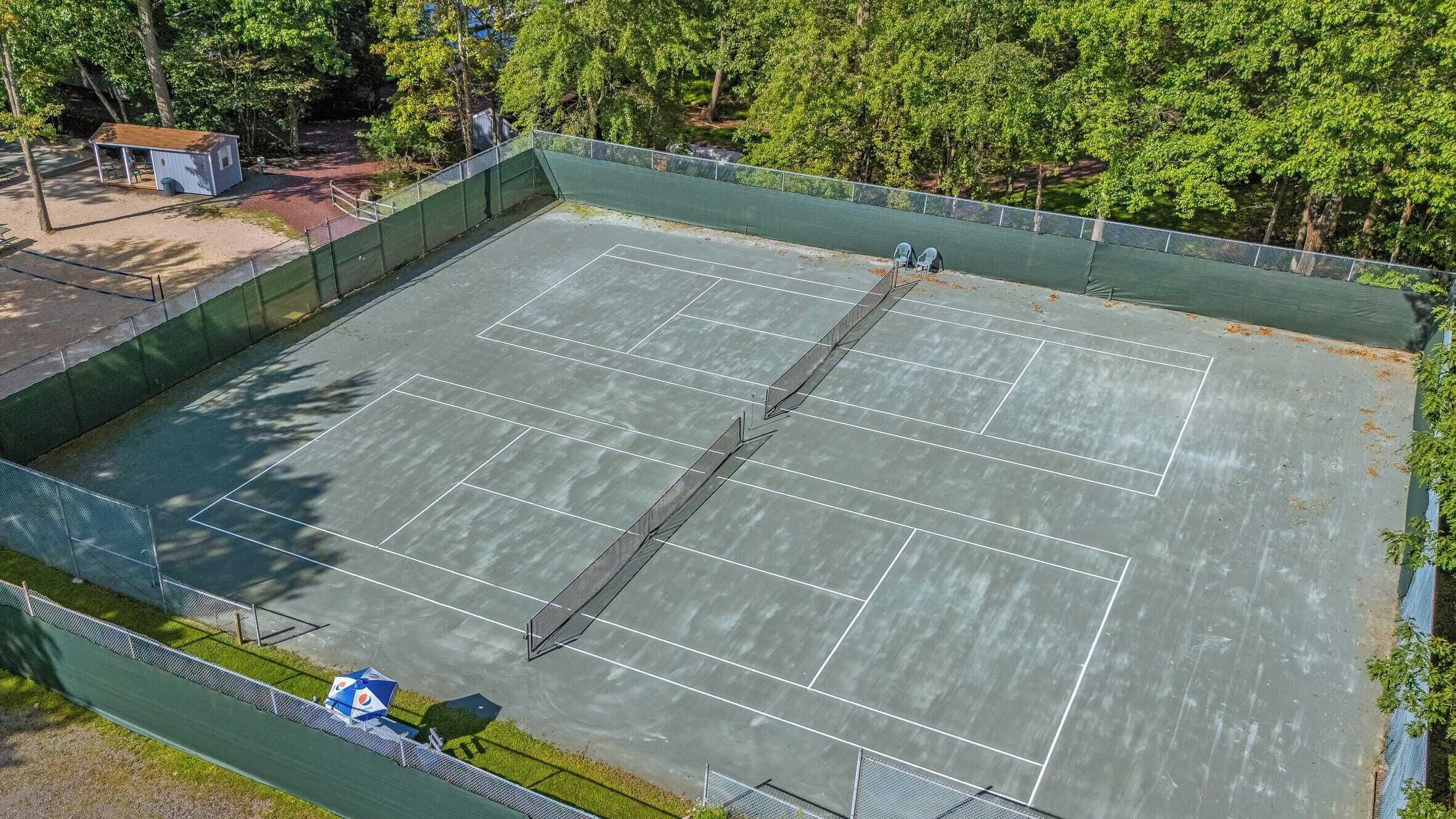 An outdoor tennis court surrounded by a green fence, with trees in the background.