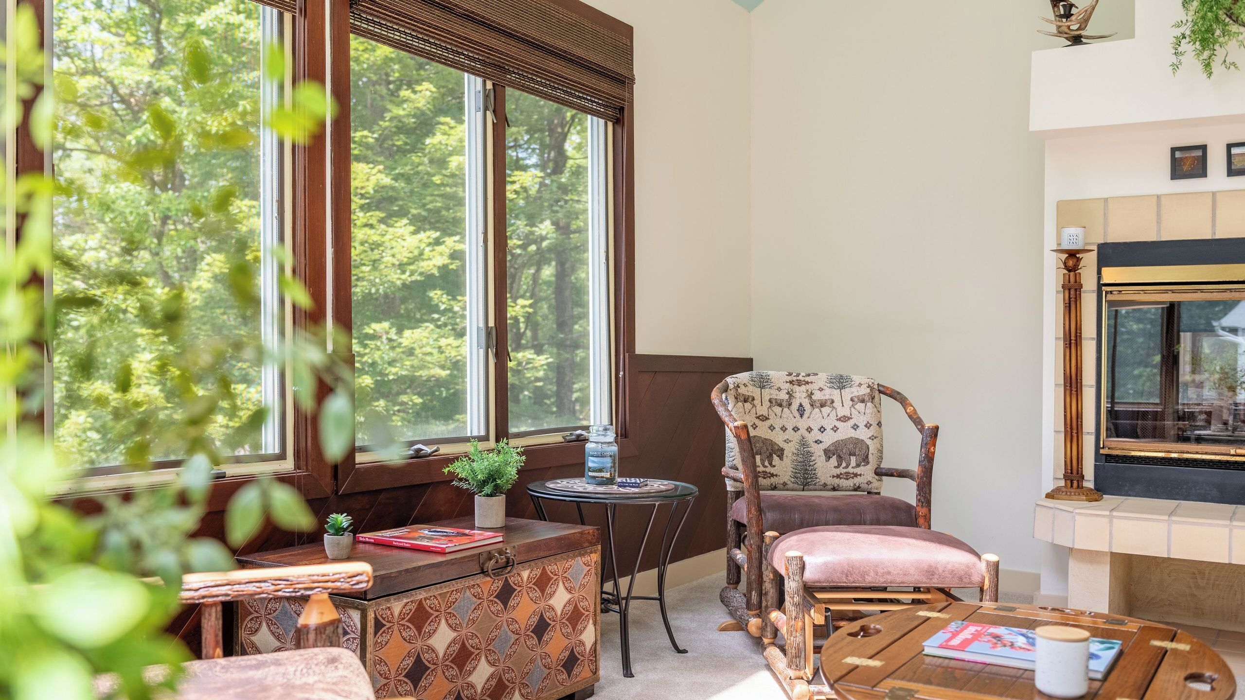 Um quarto acolhedor com uma janela grande, lareira, poltrona estampada, mesa pequena e um armário decorativo.