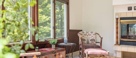 A cozy room with a large window, a fireplace, a patterned chair, a small table, and a decorative cabinet.