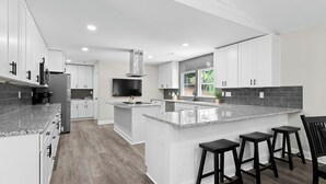 Een moderne keuken met witte kasten, granieten aanrechtbladen, een centraal eiland met barkrukken, een inbouwfornuis en een groot raam dat voor natuurlijk licht zorgt.