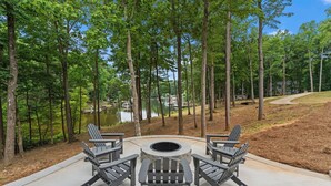 Een rond zitgedeelte buiten met Adirondack-stoelen en een vuurkuil, omringd door een bebost landschap en een meer.