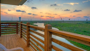 Une terrasse en bois, dotée d’une balustrade, qui offre une vue sur un champ verdoyant et une étendue d’eau, avec un coucher de soleil en arrière-plan.