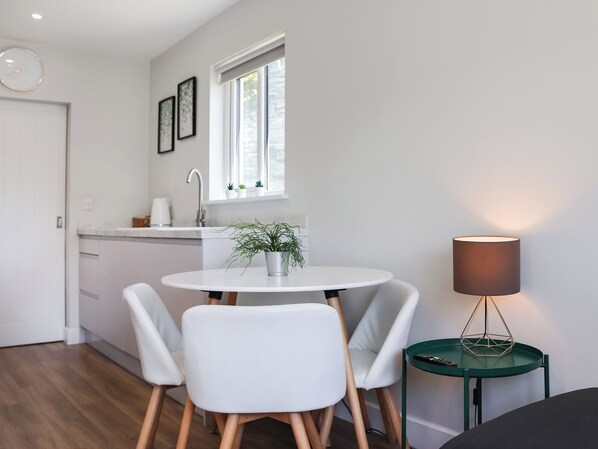 A modern kitchen with white cabinets, a white countertop, and a window above the sink. There is a round white table with two chairs and a small green side table with a lamp.