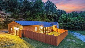 A wooden cabin with a fenced backyard, a covered porch, and a well-lit interior.