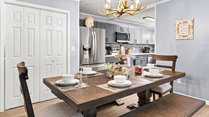 A dining room with a wooden table set for four, white chairs, a chandelier, and a kitchen in the background.