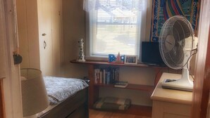 A compact bedroom with a single bed, a desk with a computer, a standing fan, a window with blinds, and a decorative wall hanging. - Central air, dog friendly walk to beach in this 2 bedroom family cottage  (Dennis)