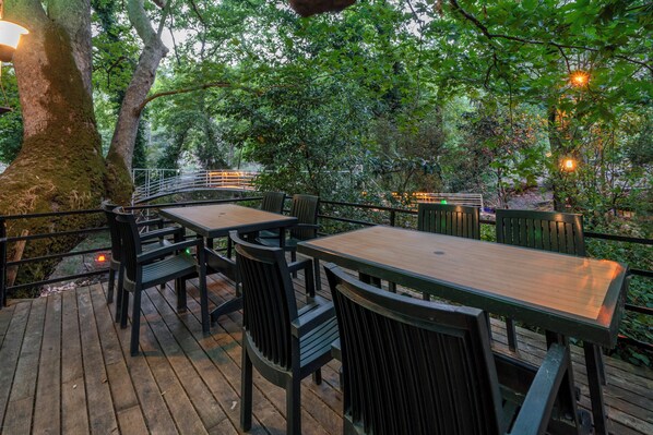 An outdoor wooden deck with tables and chairs, surrounded by trees and a railing. - Yoruk Park Pansiyon (Kemer)