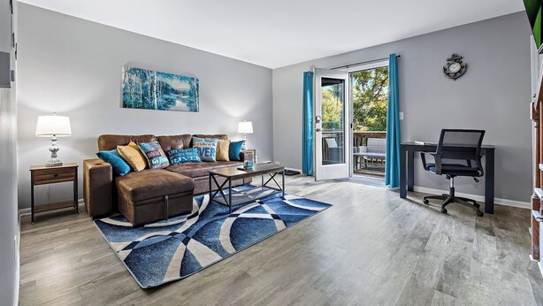 A modern living room with a sofa, coffee table, and a desk with a chair. There is a painting on the wall and a view of the outdoors through a glass door. - Relaxing 2BR Riverfront Townhome (Yorkville)