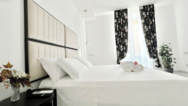 A neatly made bed with white linens and pillows, a bedside table with a remote control, a vase with flowers, and a potted plant.