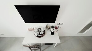 A white table with a flat-screen TV, a coffee maker, a vase with flowers, and a tray of white teacups and saucers.