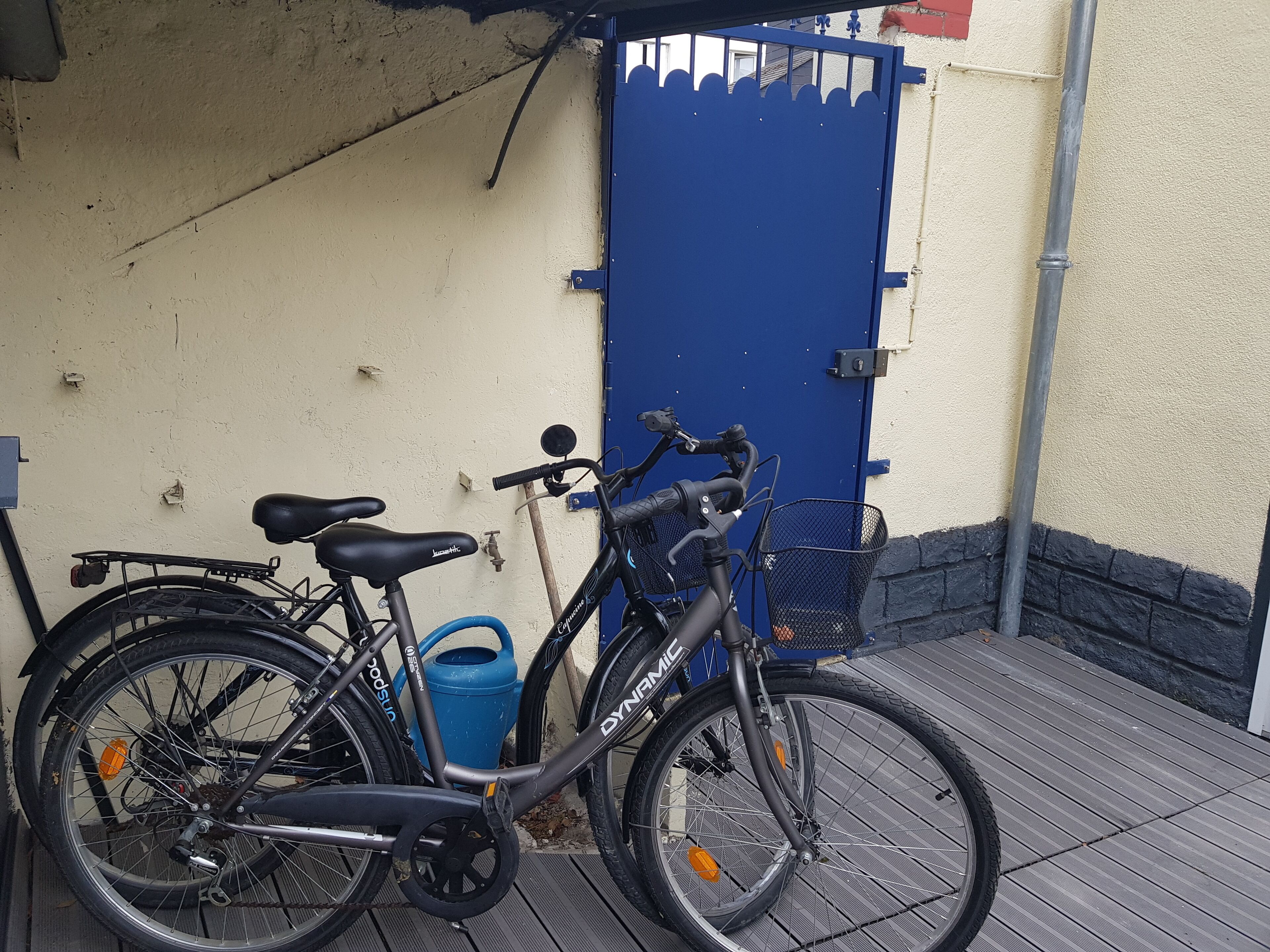 A bicycle parked next to a blue gate, with a blue trash bin and a water tap nearby.