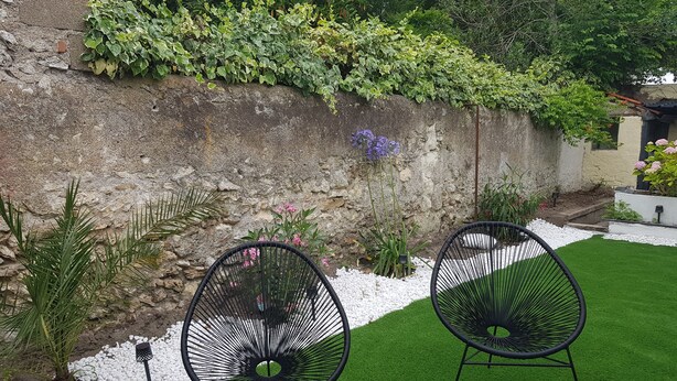 Two black wireframe chairs on a green lawn with white pebbles, a stone wall with climbing plants, and a small garden area with flowers and plants. - Cosy town house with bicycles available (Tours)