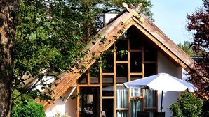 Een houten huis met een schuin dak en grote ramen. Er ligt een gazon ervoor en een tafel met stoelen onder een parasol.