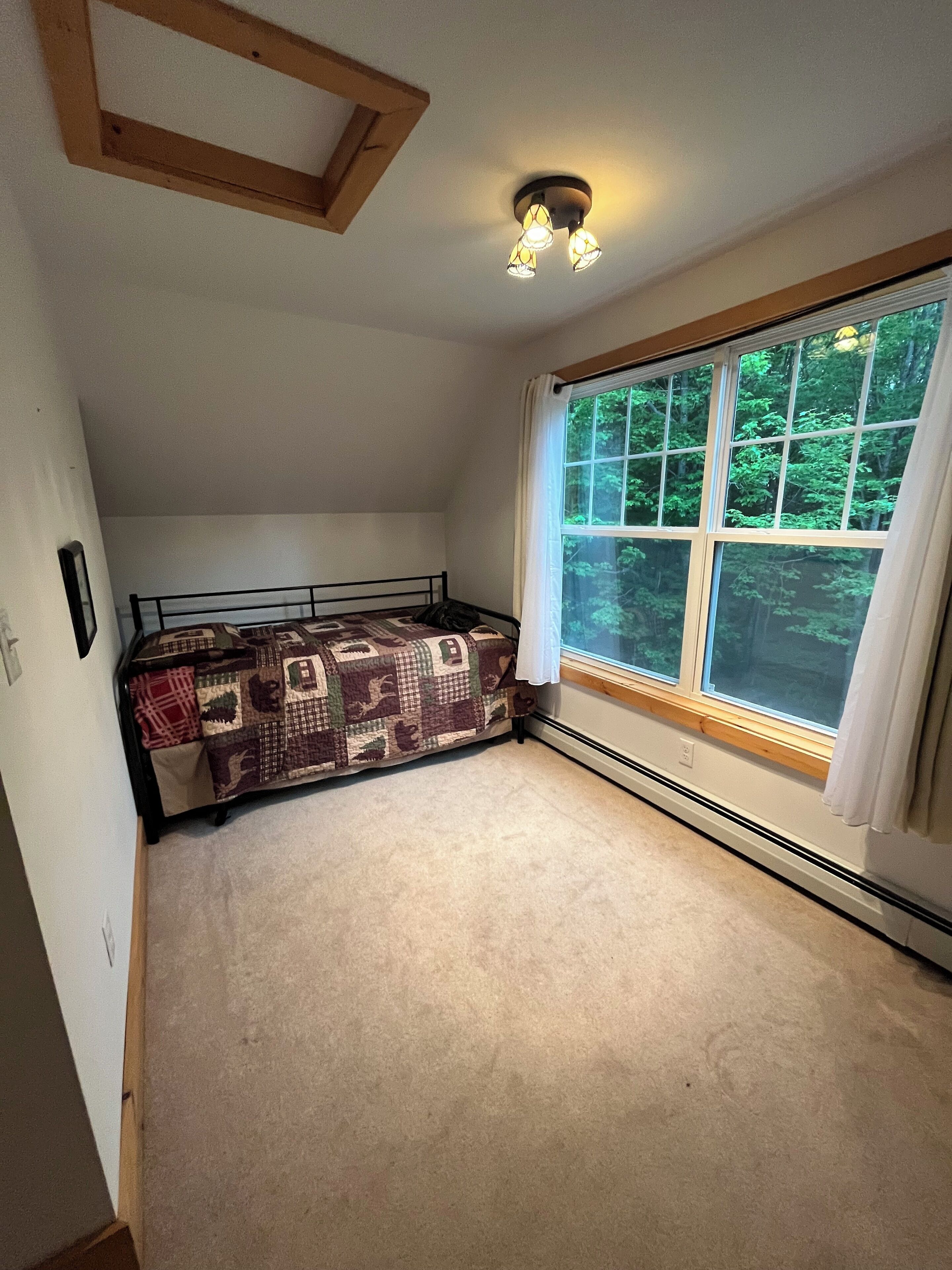 A bedroom with a sloped ceiling, a bed with a patterned quilt, a window with a view of trees, and a ceiling light fixture.