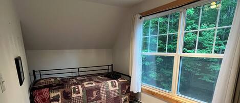A bedroom with a sloped ceiling, a bed with a patterned quilt, a window with a view of trees, and a ceiling light fixture.