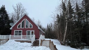 Eine rote Holzhütte mit weißem Vorbau und Stufen zu einer Terrasse, umgeben von Schnee und Bäumen.