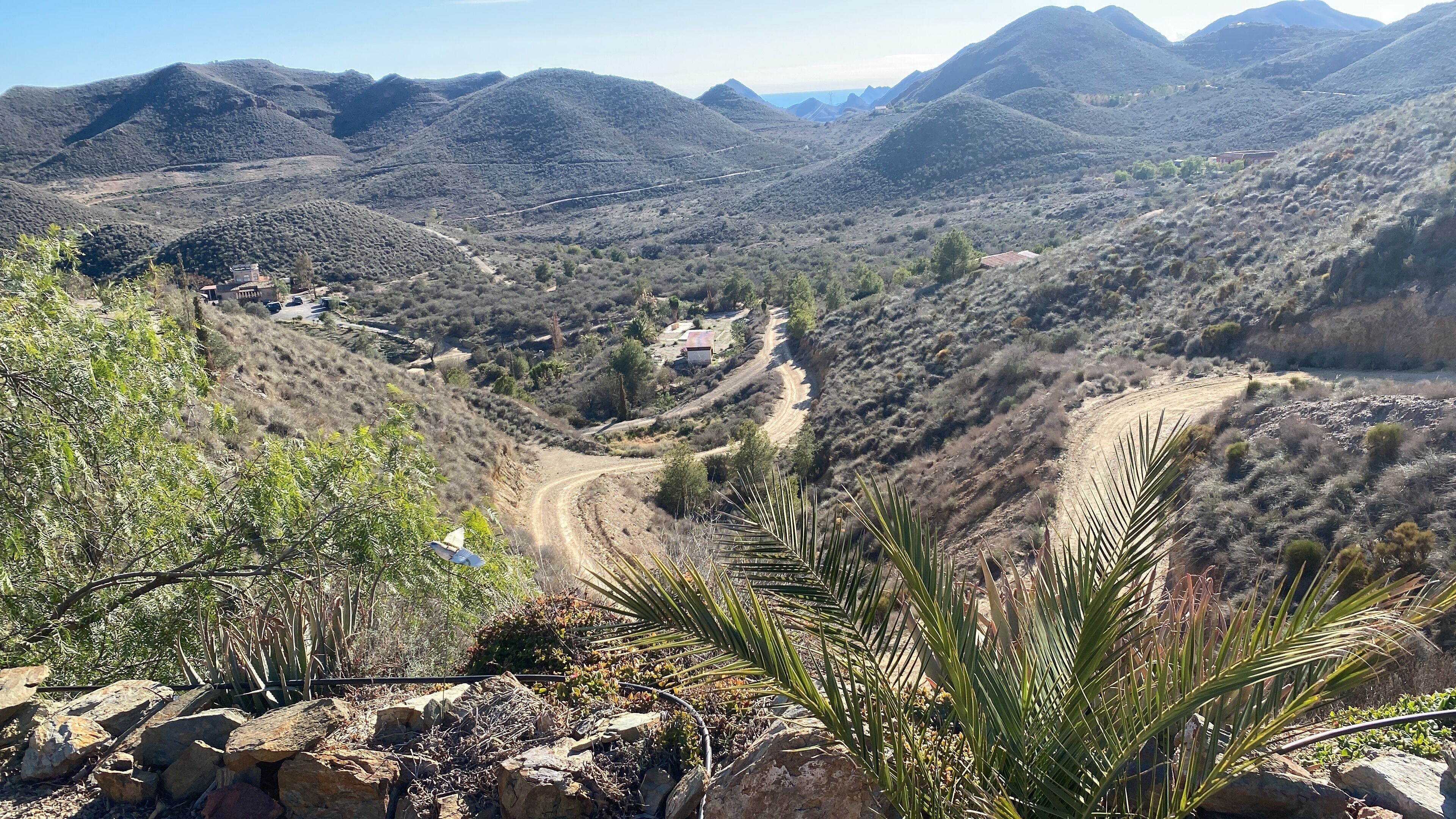 A mountainous landscape with a winding dirt road, sparse vegetation, and a few buildings in the distance.