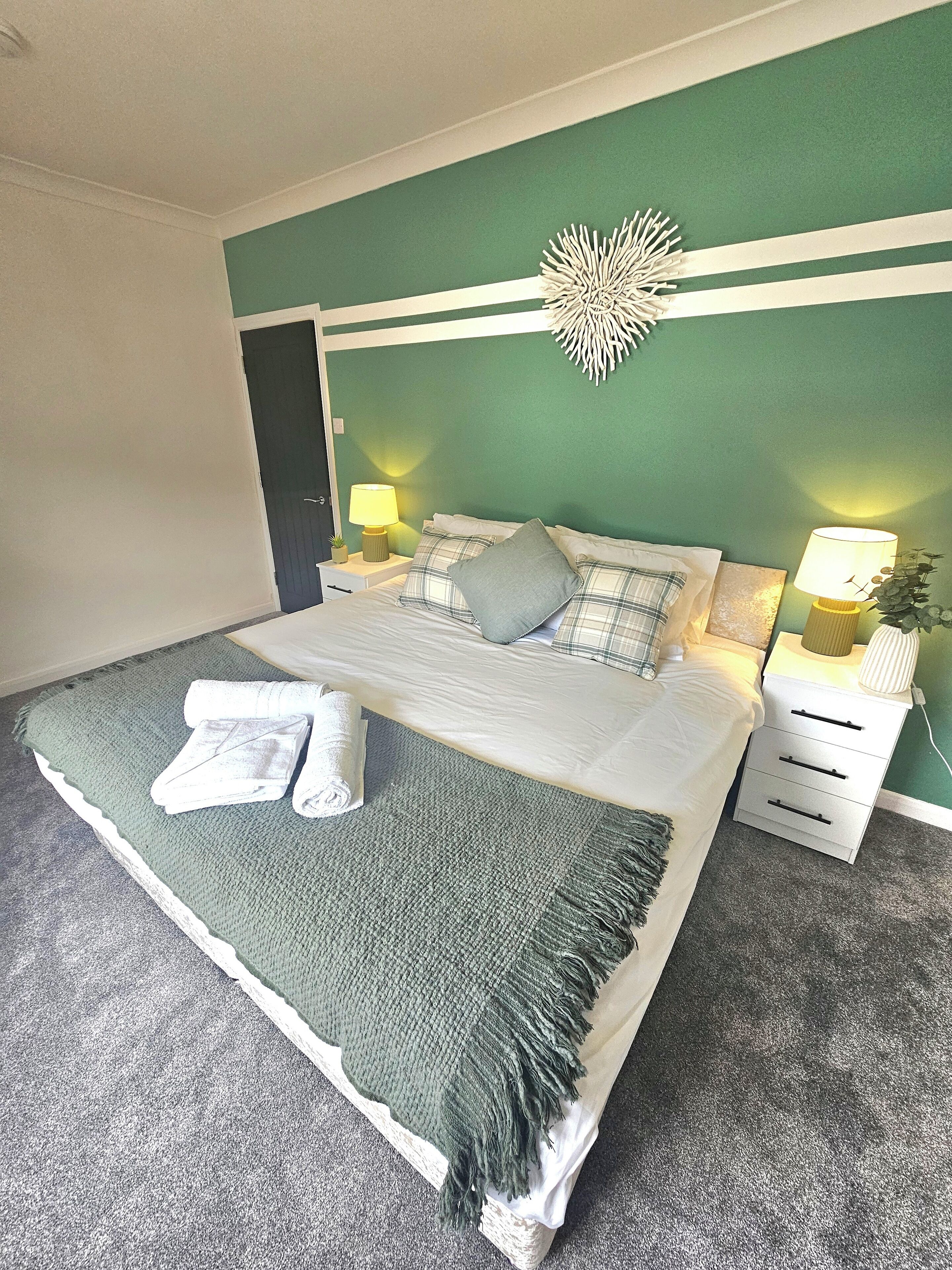A neatly made bed with a green blanket and pillows, a bedside lamp, and a decorative wall piece.