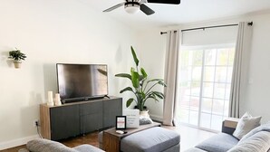 A modern living room with a grey sofa set, a flat-screen TV on a wooden cabinet, a potted plant, and a sliding glass door leading to an outdoor area. - Resort Style Home Near Universal/Disney (Orlando)