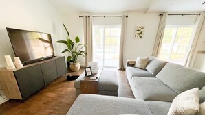 A modern living room with a grey sofa, a flat-screen TV on a wooden cabinet, a potted plant, and a small table with a remote control. - Resort Style Home Near Universal/Disney (Orlando)