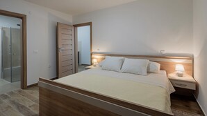A modern hotel room with a wooden bed, bedside tables with lamps, a glass shower enclosure, and a white ceiling light. - Holiday Home Milutin 1 - Four Bedroom Holiday Home with Private Pool (Vrsi)