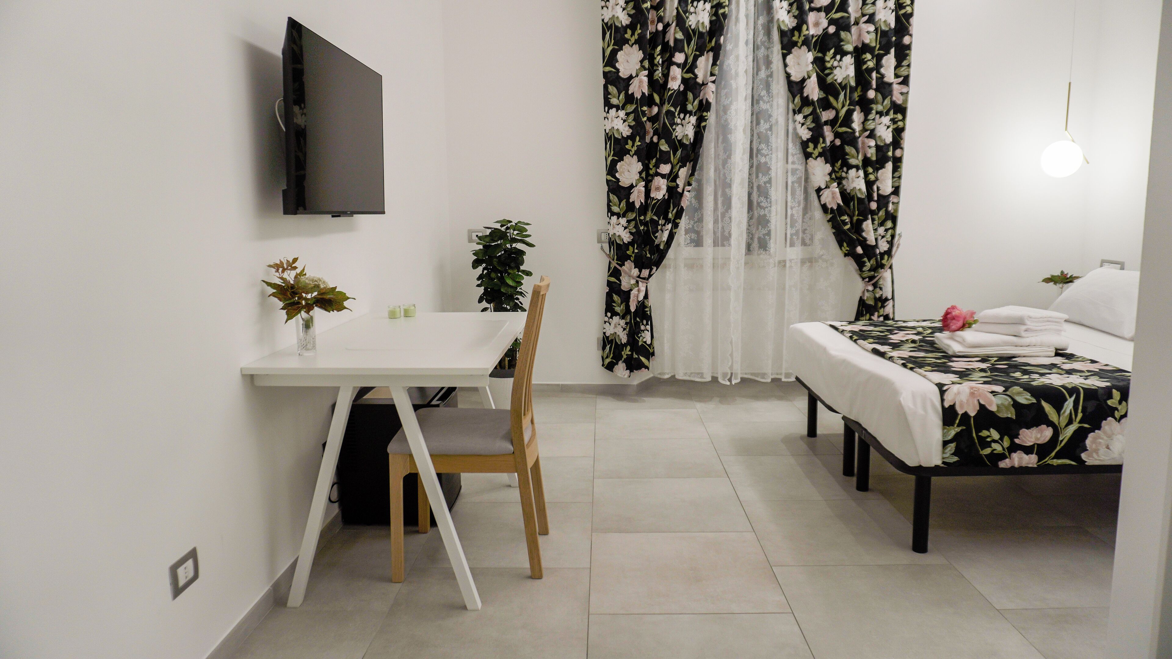 A compact room with a bed, a desk, and a chair. There is a TV mounted on the wall, a plant on the desk, and a window with floral curtains.