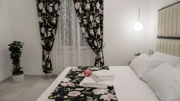 Una cama bien hecha con sábanas blancas, una colcha de estampado floral y una sola flor rosa. Hay una planta en maceta a la izquierda y una mesita de noche con una lámpara.