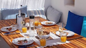 Une table en bois, dressée avec des assiettes, des verres et une bouteille, entourée de lampes suspendues en osier et d’un mur blanc orné d’un pot en pierre.