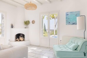 A bright living room with a fireplace, a sofa, a floor lamp, and a view of the outdoors through a glass door. - Sea-View Cycladic Villa near Alyki – Family Home with Olive Garden & Starlink (Paros)