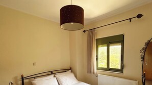 A neatly made bed with white linens and a folded towel on the side. There is a bedside table with a lamp and a small box, possibly for tissues. A window with a curtain is visible, and a radiator is mounted on the wall.
