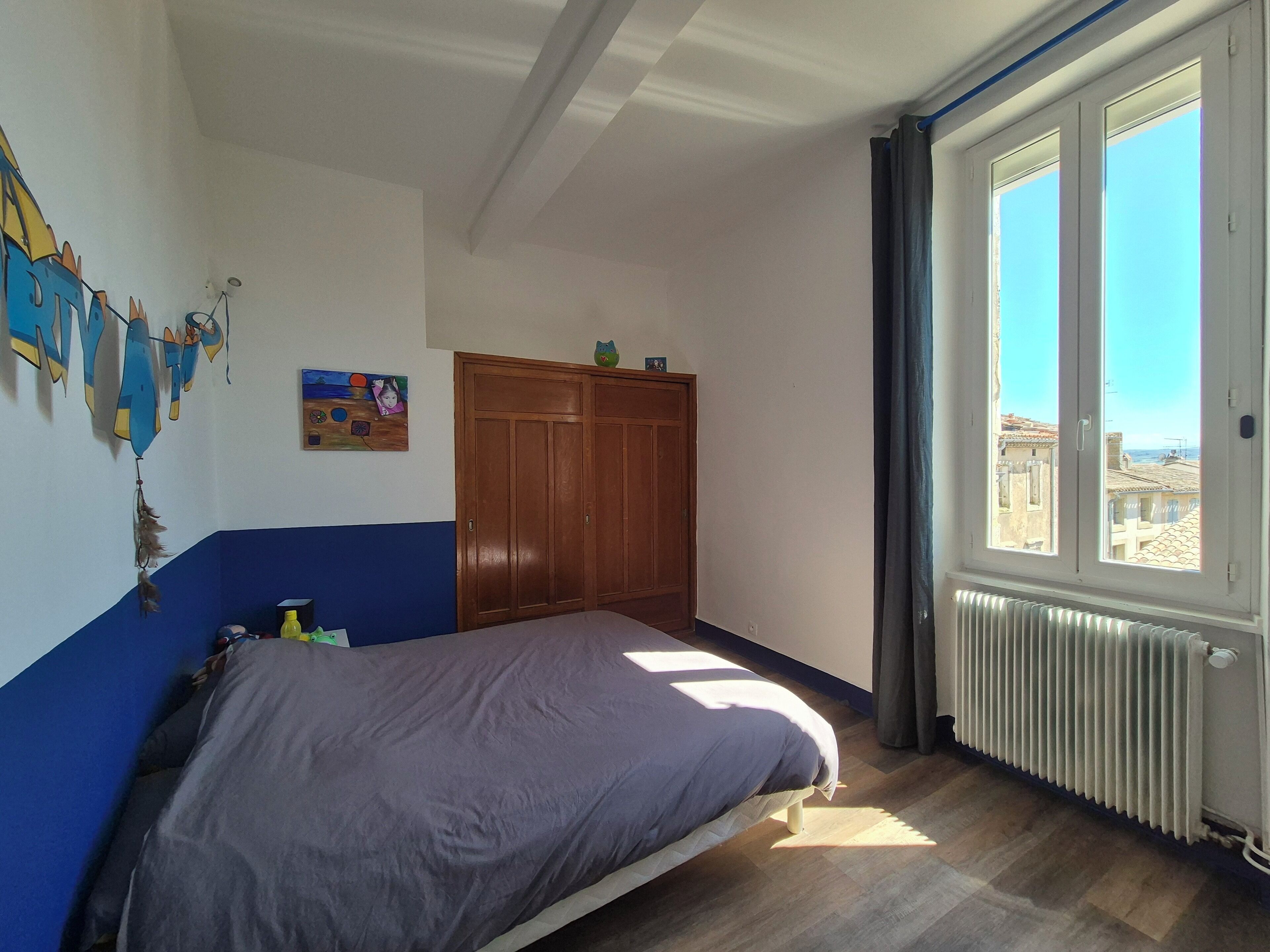 A bedroom with a bed, a wardrobe, a radiator, and a window with a view of buildings.