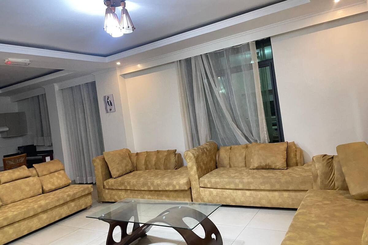 A modern living room with beige sofas, a glass coffee table, and sheer curtains.
