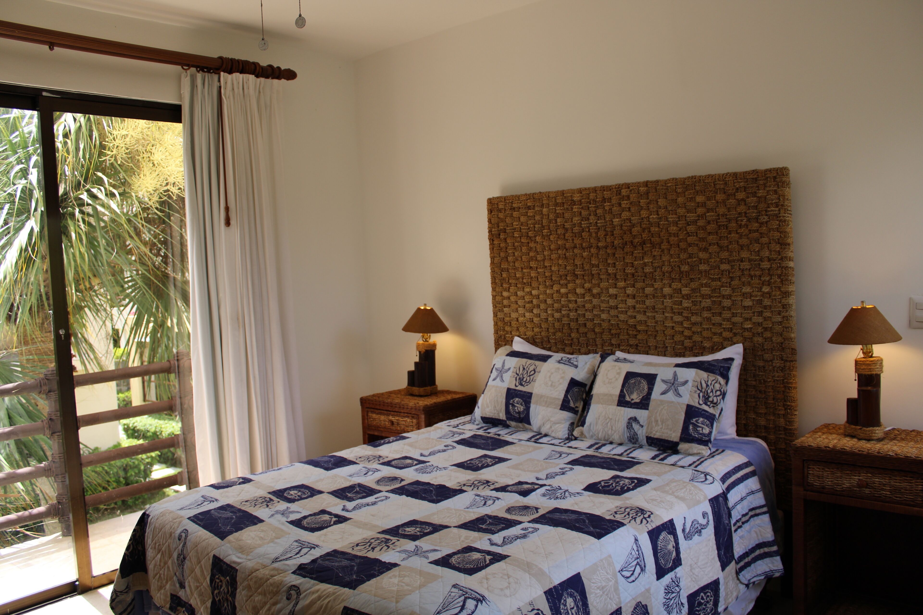 A bedroom with a large bed, two bedside tables with lamps, a woven headboard, and a view of palm trees through a window with a curtain.