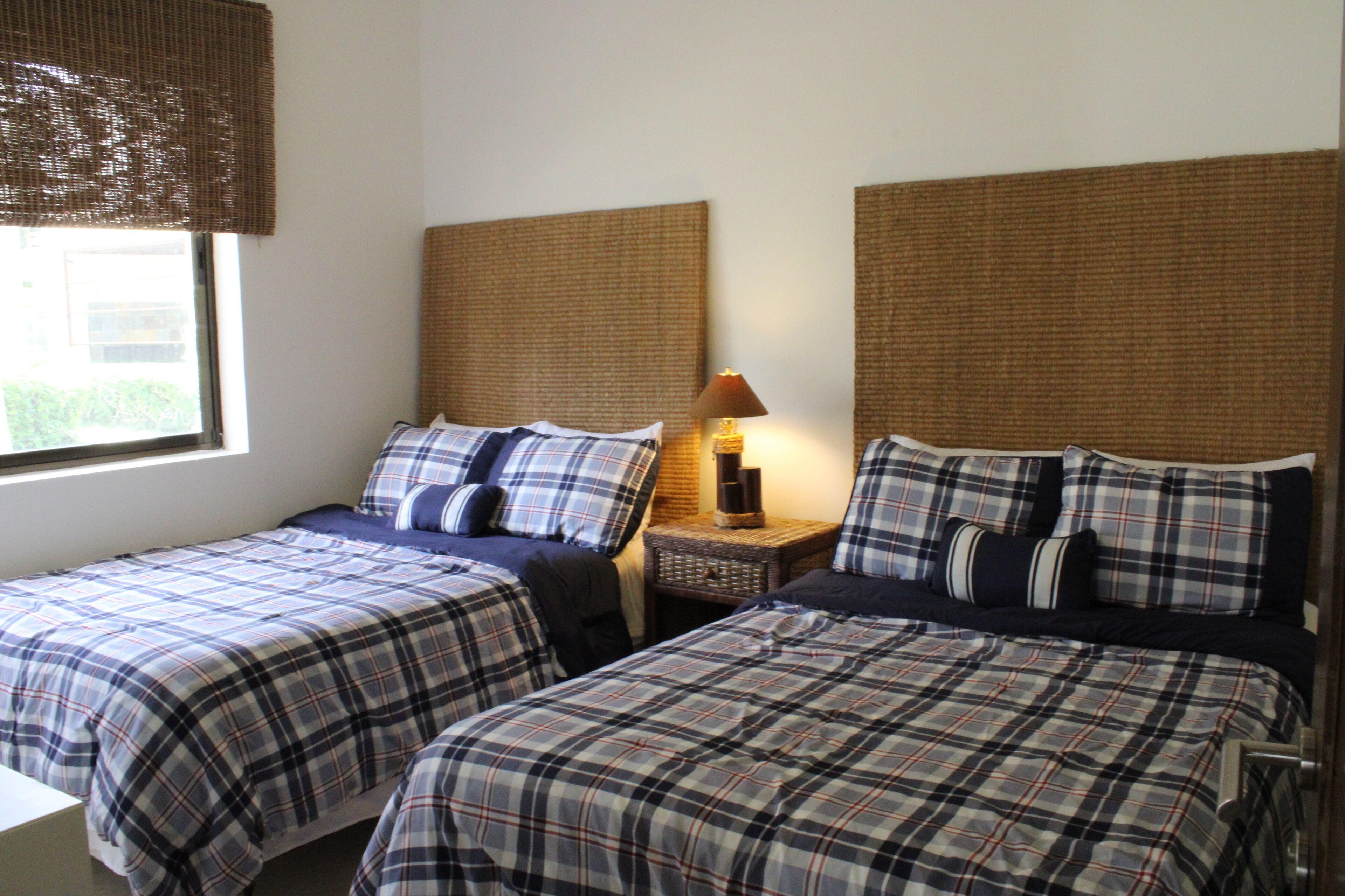 A room with two beds, each with a plaid bedspread and matching pillows. There is a nightstand with a lamp between the beds.