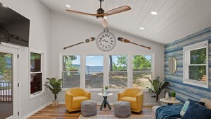 Una habitación amplia con un ventilador de techo grande, un reloj de pared redondo y vistas al exterior a través de amplias ventanas.