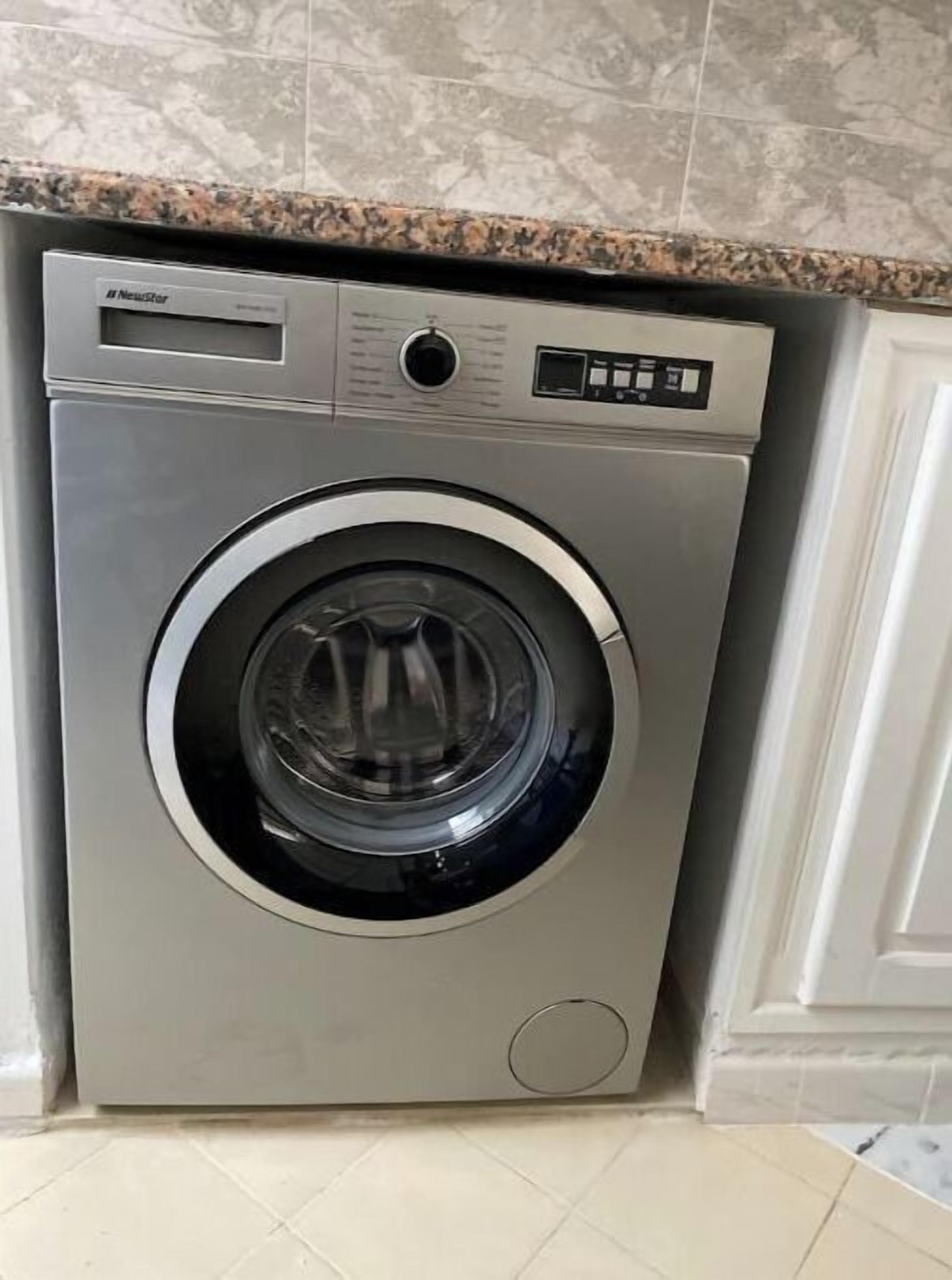 Una lavatrice a carica frontale compatta, in acciaio inossidabile, con pannello di controllo digitale, posizionata in una cucina con piano di lavoro in granito e mobili bianchi.